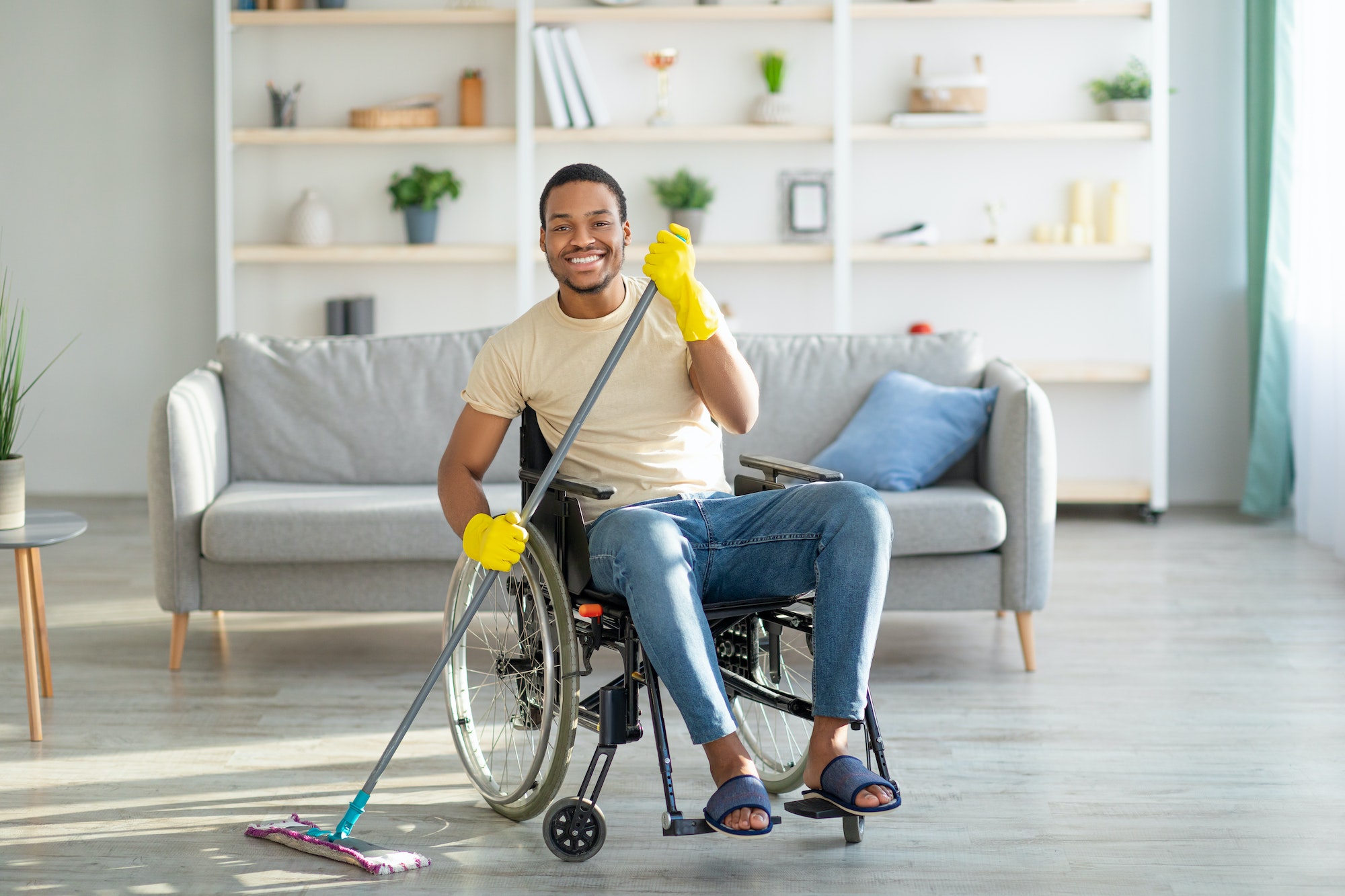 Disabled black man in wheelchair washing floor with mop at home. House cleanup concept